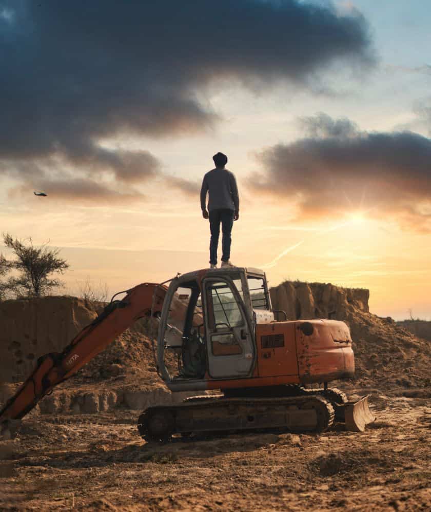A man on an excavator at sunset, with the sky painted in hues of orange and purple.